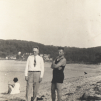 Two men standing on beach, one in swimming suit, one wearing a tie.