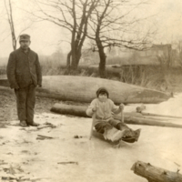 9-15-v: Penobscot boy on sled, man standing behind, canoe in background.