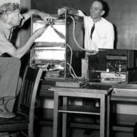 W. F. G. Swann, foot on chair, working on equipment with unidentified man.