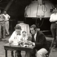 Two men, seated at table with NBC microphone, gondola for stratosphere flight in background.