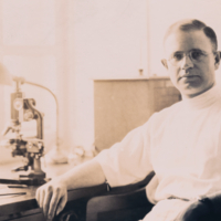 Harold Amoss, half length, informal, sitting in lab at desk with microscope.