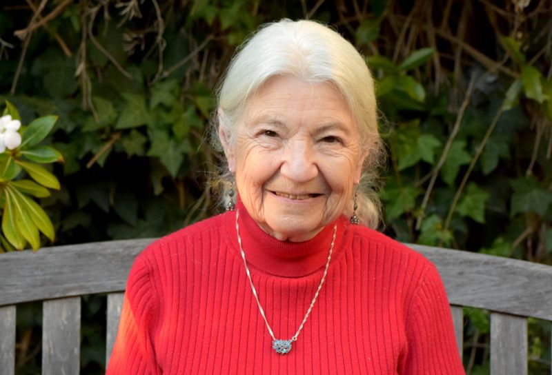 Portrait of Baycsy dressed in a red sweater and seated in front of a wooden fence with leafy plants behind her.