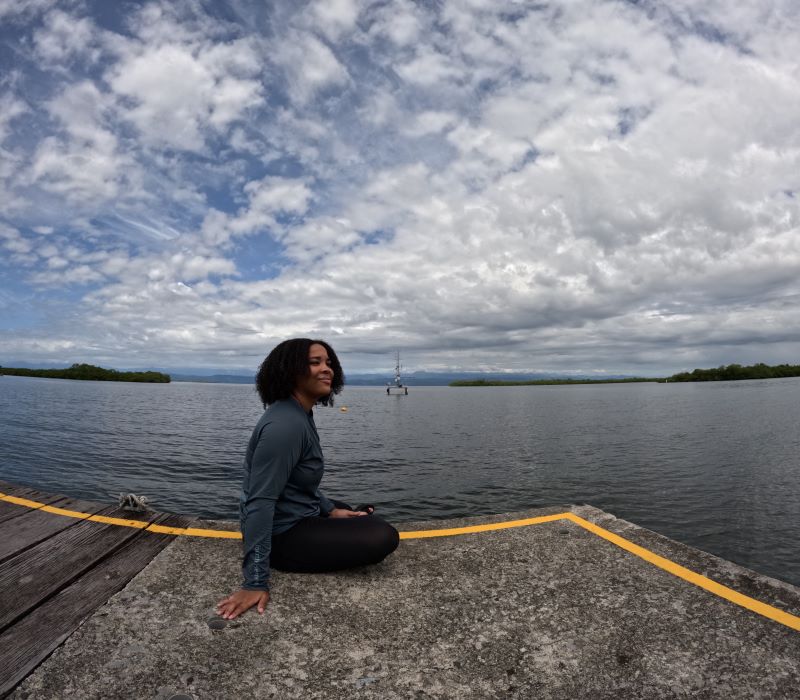 Boyd, a woman with curly black hair, is seated on a dock in front of a placid body of water, dressed in a casual outfit.