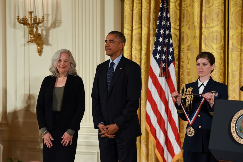 Judith Klinman standing next to President Barack Obama while a government official in uniform presents the National Medal of Science.