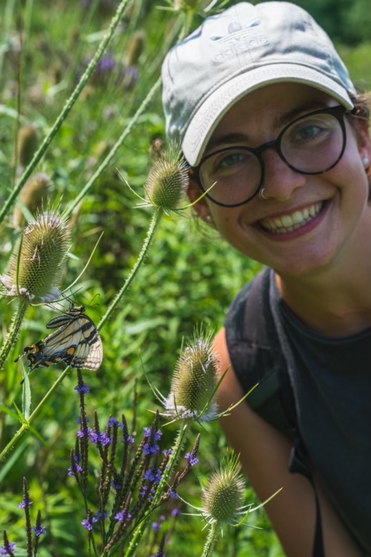 Catherine McManus smiling next to a butterfly perched on a plant.