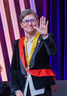 Deb Niemeier wearing a sash smiling and waving at the camera.