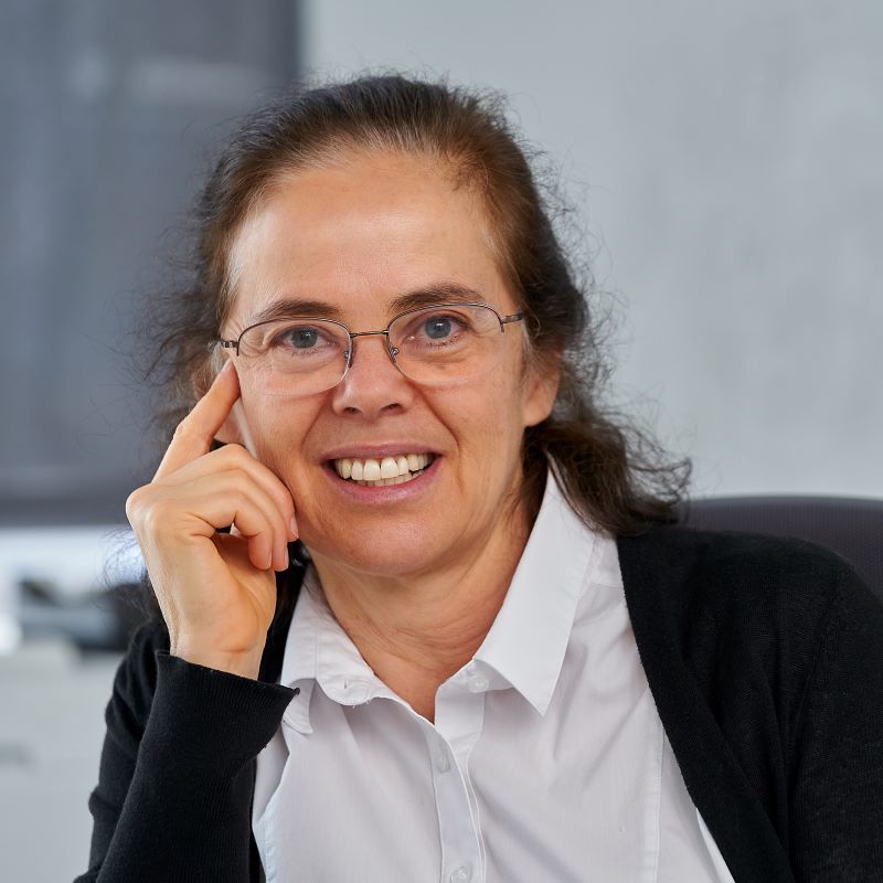 Close-up image of a woman with glasses wearing a white blouse and black cardigan, her hand resting on the side of her face with her pointer-finger extended upward. 